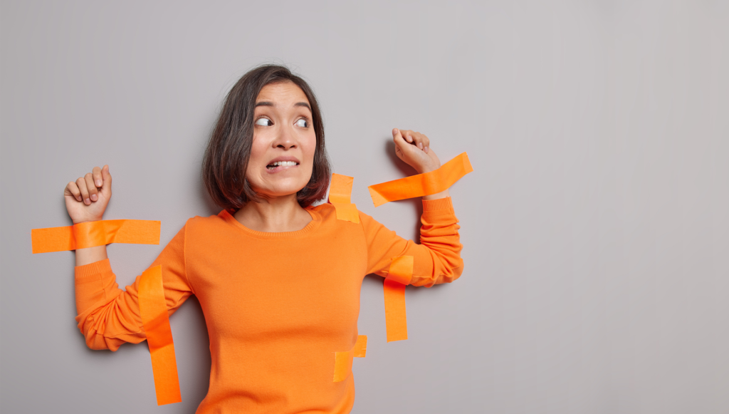 Frightened woman with dark hair stuck with adhesive tapes to grey wall bites lips has nervous expression isolated